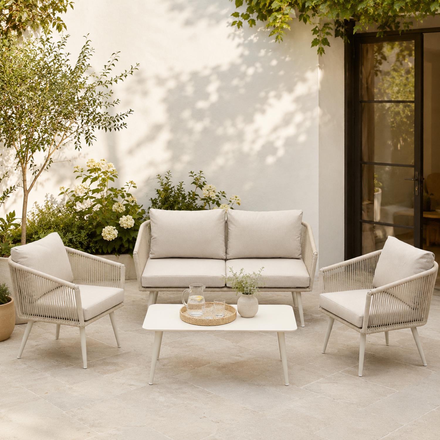 Outdoor patio set with beige sofa, chairs, and table on a patio with plants and a white wall.